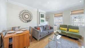 A modern living room with a dining area, a green sofa, and a city view. - Cozy 2-Bedroom Loft next to Park Lane (London)