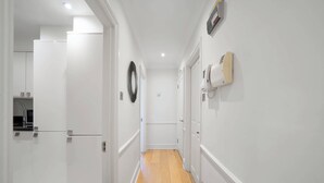 A narrow hallway with white walls, a wooden floor, and a door at the end leading to a kitchen area. - Cozy 2-Bedroom Loft next to Park Lane (London)