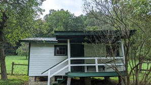Une petite cabane à un seul étage, dotée d’un toit vert et de murs blancs, avec un porche à l’avant et un escalier menant à une porte.