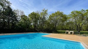 A clear blue swimming pool surrounded by trees and a paved area with a bench. - Chalet Nature - Lodges du Bois Dodo (Septfonds)