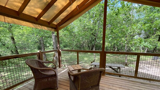 Une terrasse en bois couverte, dotée de chaises en osier et d’une table, donnant sur un espace clos planté d’arbres.
