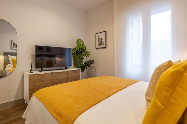 A bedroom with a bed, a TV on a wooden dresser, a large plant, and a window with curtains. - Charming Studio in Chamberí, Madrid (Madrid)