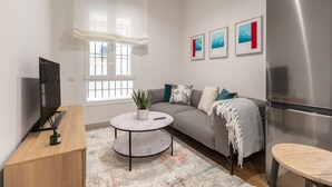 A modern living room with a grey sofa, a round coffee table, a TV on a wooden stand, a refrigerator, and a window with white shutters.