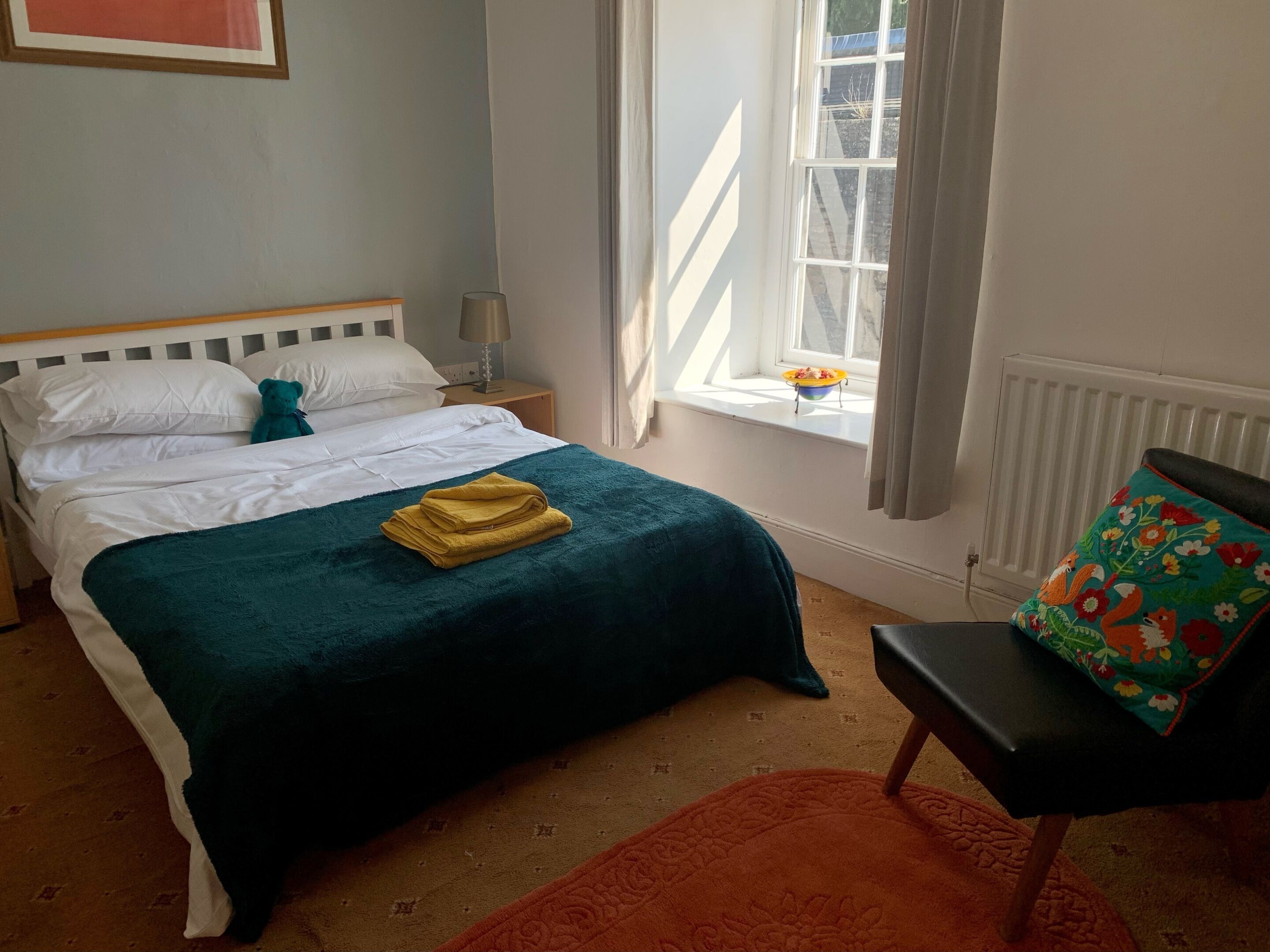 Een keurig opgemaakt bed met witte lakens en een blauwgroene deken, een nachtkastje met een lamp, een raam met gordijnen en een kleurrijke stoel.