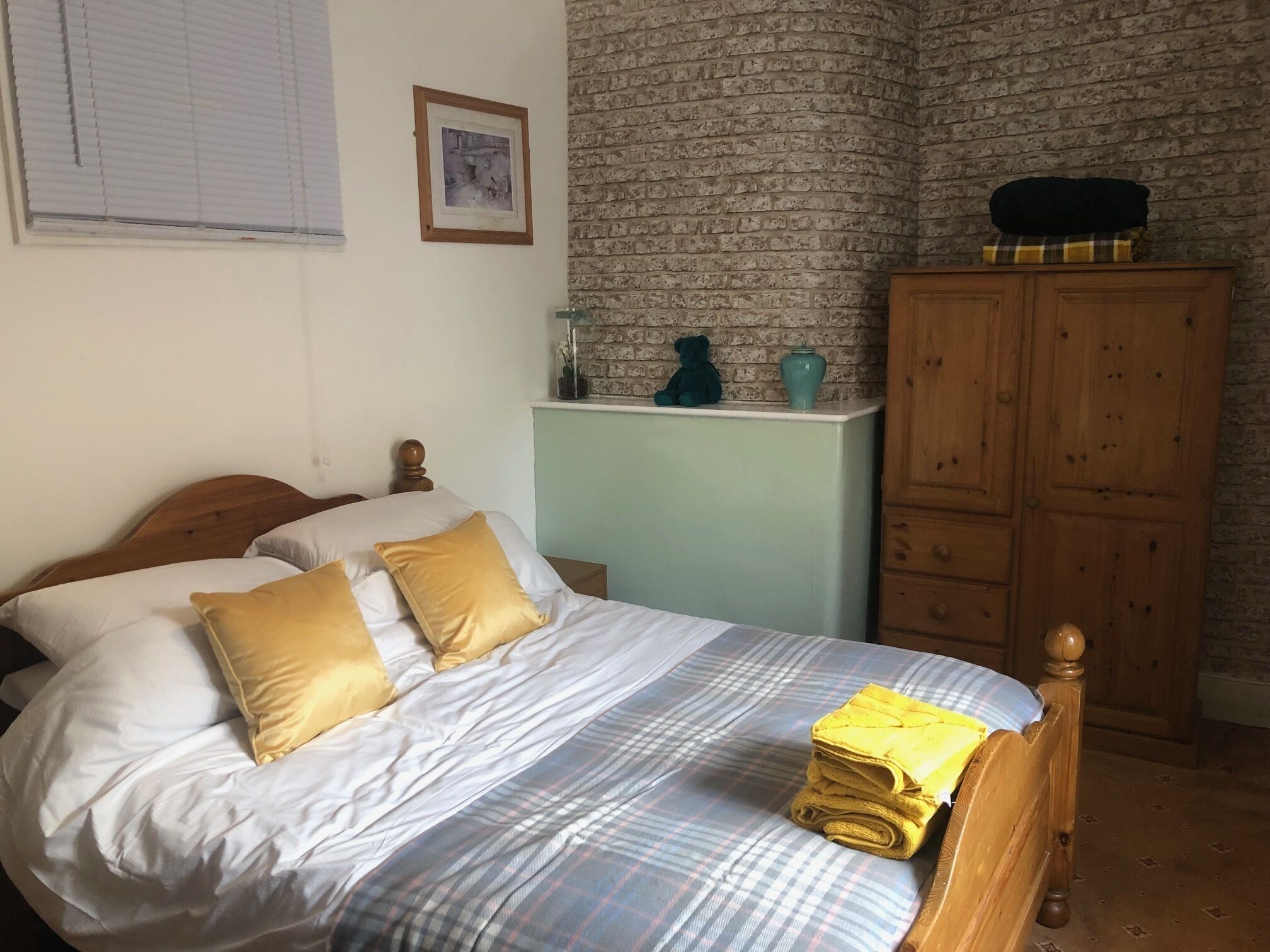 Ein ordentlich bezogenes Bett mit weißer und goldener Bettwäsche, einer Karo-Decke und einem Stapel gelber Handtücher. Im Hintergrund ist ein hölzerner Kleiderschrank und eine Wand mit Ziegelmuster zu sehen.