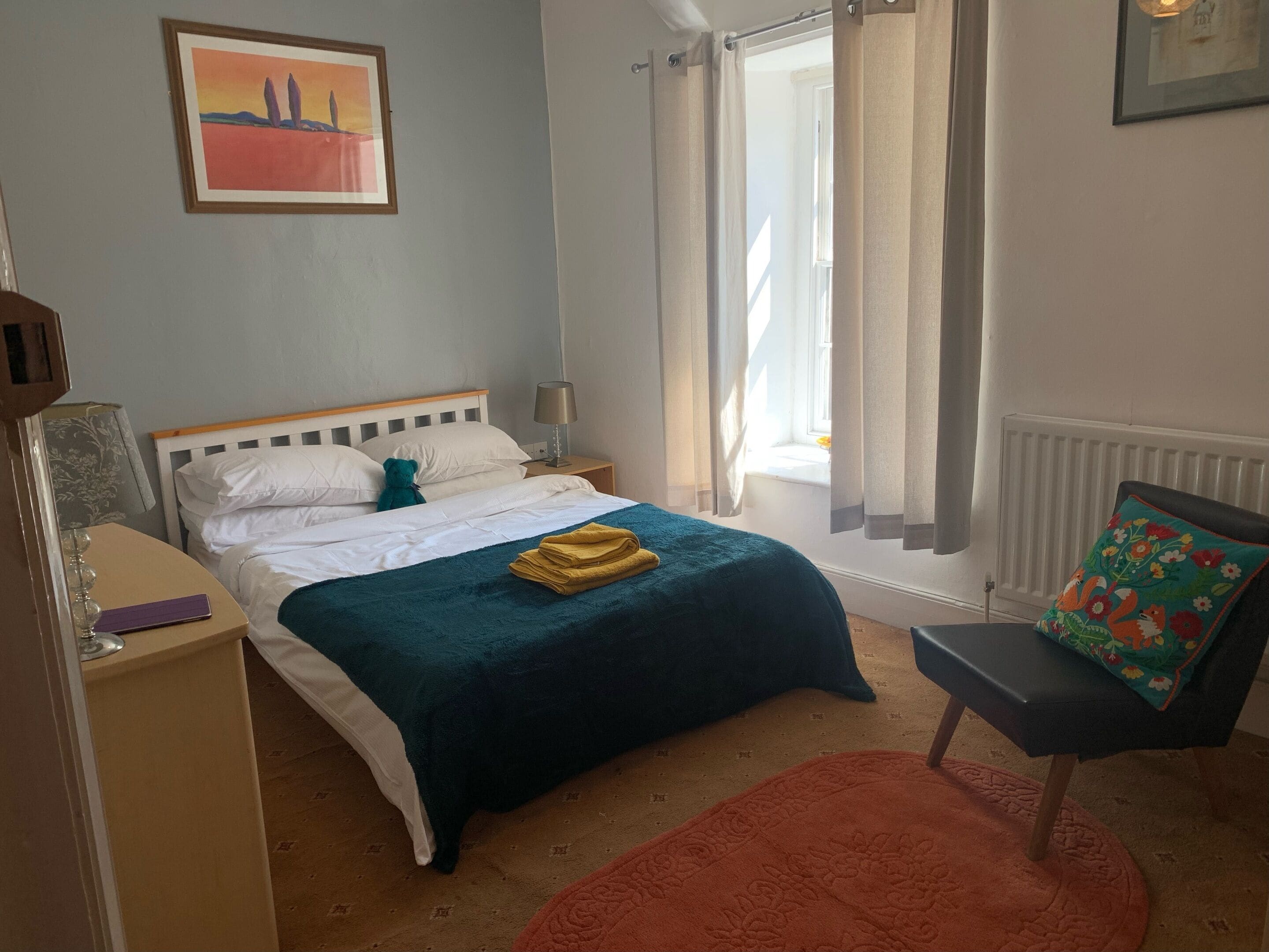 Un dormitorio con cama, mesita de noche, silla, ventana con cortinas y radiador.