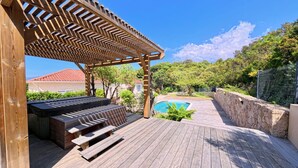 Una terraza de madera con pérgola, piscina y un muro de piedra.
