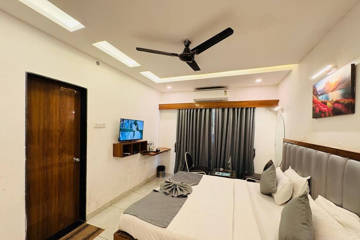 A hotel room with a bed, a ceiling fan, a TV, a window with curtains, and a painting on the wall.