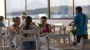 A dining area with wooden chairs and tables, large windows with a view of the outdoors, and a waiter serving food.