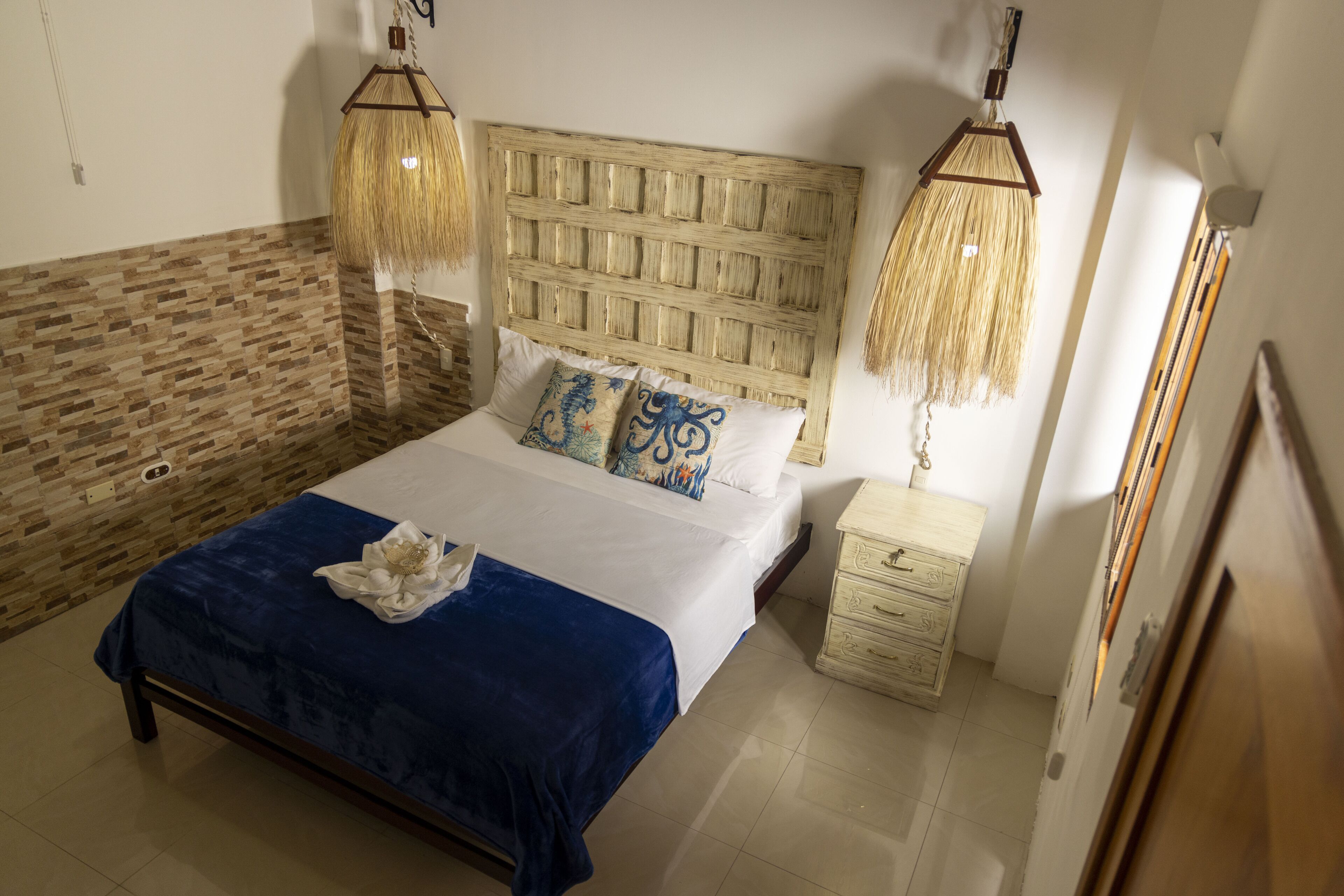 Ein Schlafzimmer mit einem hölzernen Kopfteil, einer blauen Tagesdecke, dekorativen Kissen, einem Nachttisch und einer Pendelleuchte.