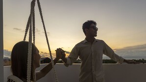 A couple holding hands on a rooftop terrace with a sunset in the background.
