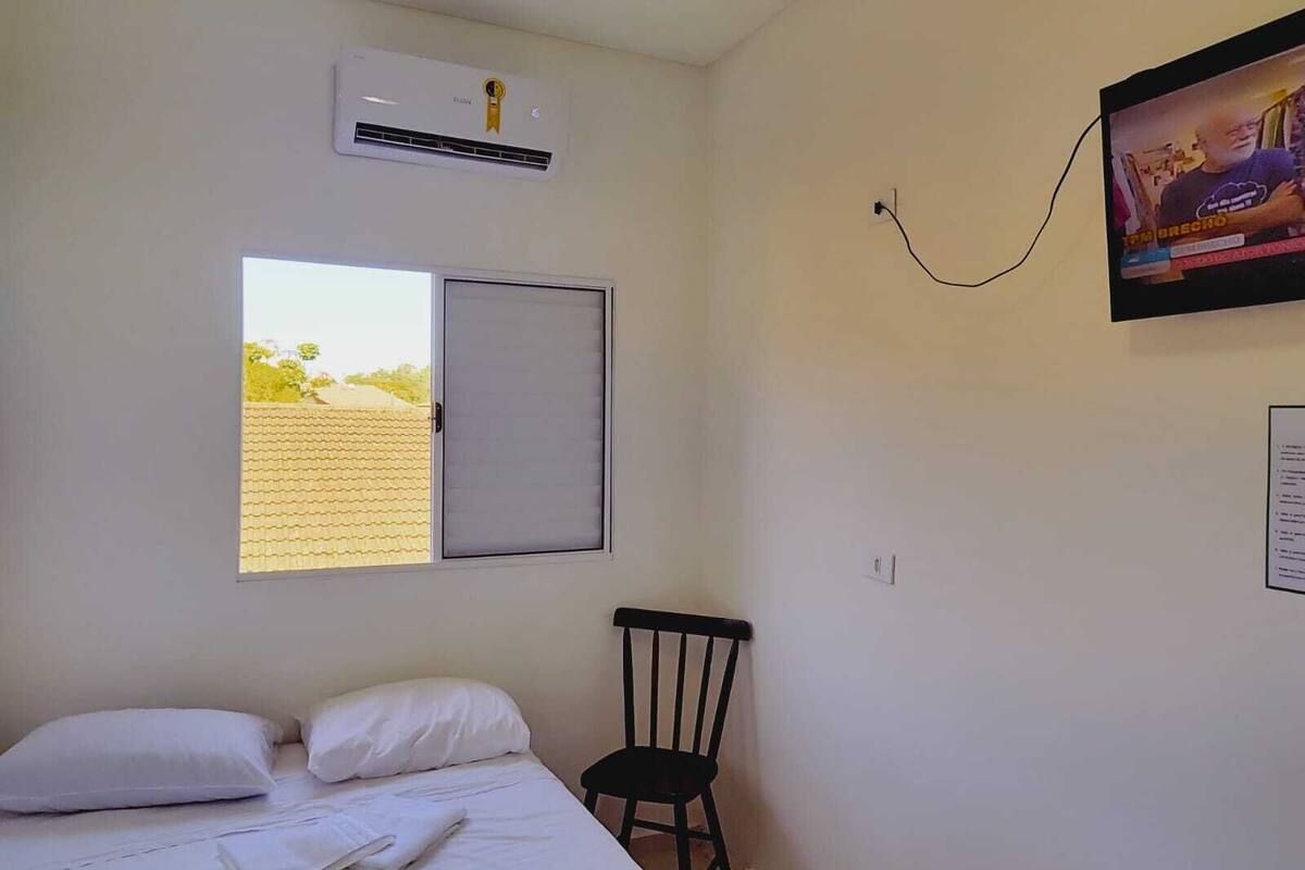 A bedroom with a bed, a chair, an air conditioning unit, a window, and a wall-mounted TV.