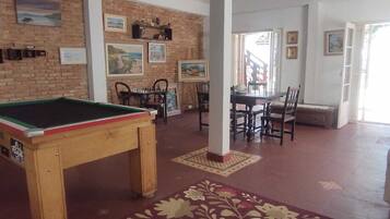 A billiards table in a room with a brick wall, several framed pictures, and a dining area with chairs and a table.