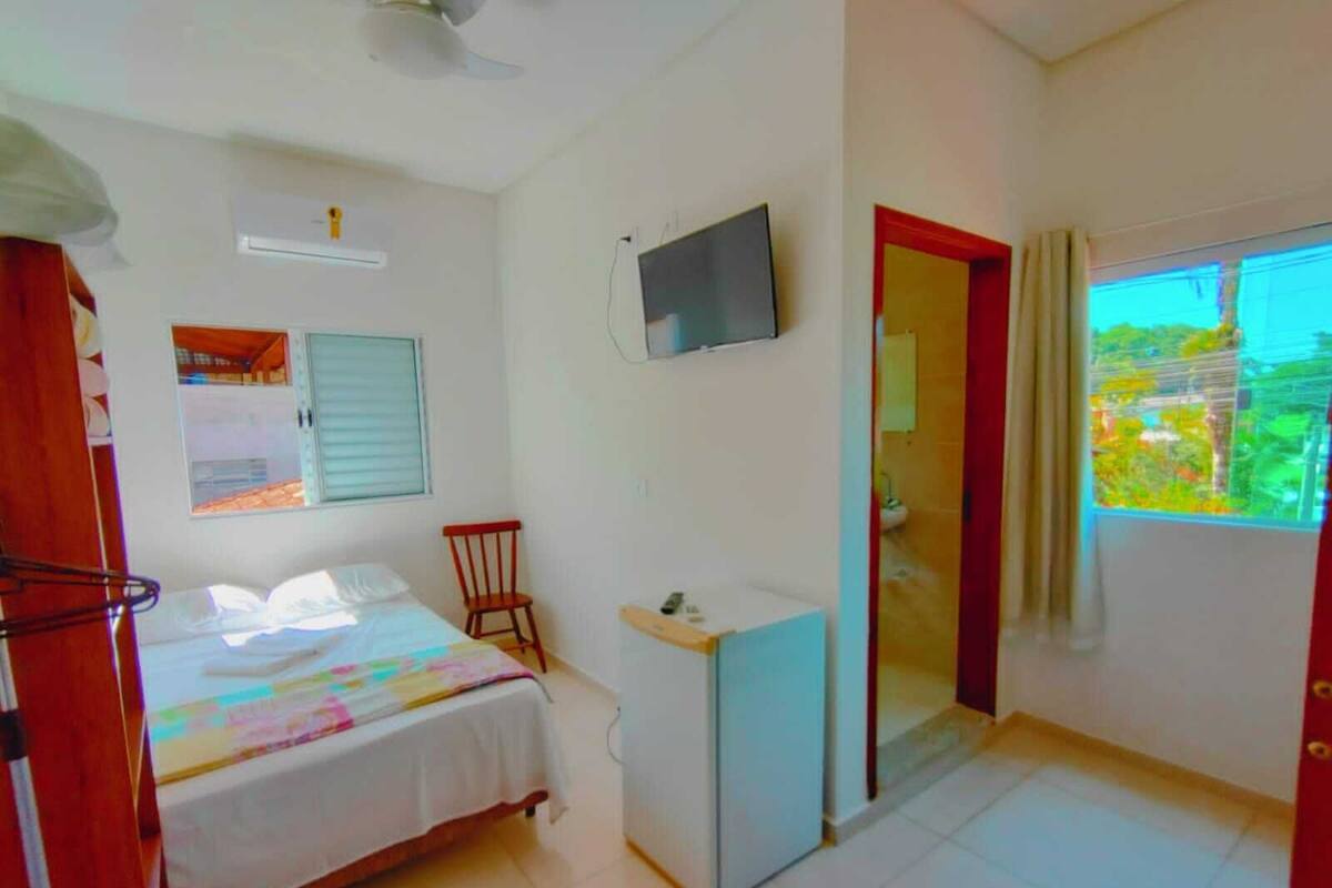 A bedroom with a bed, a desk, a chair, a TV, and a window with a view of greenery.