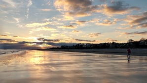 Una playa con una persona caminando sobre la arena, casas al fondo y un atardecer en el cielo.