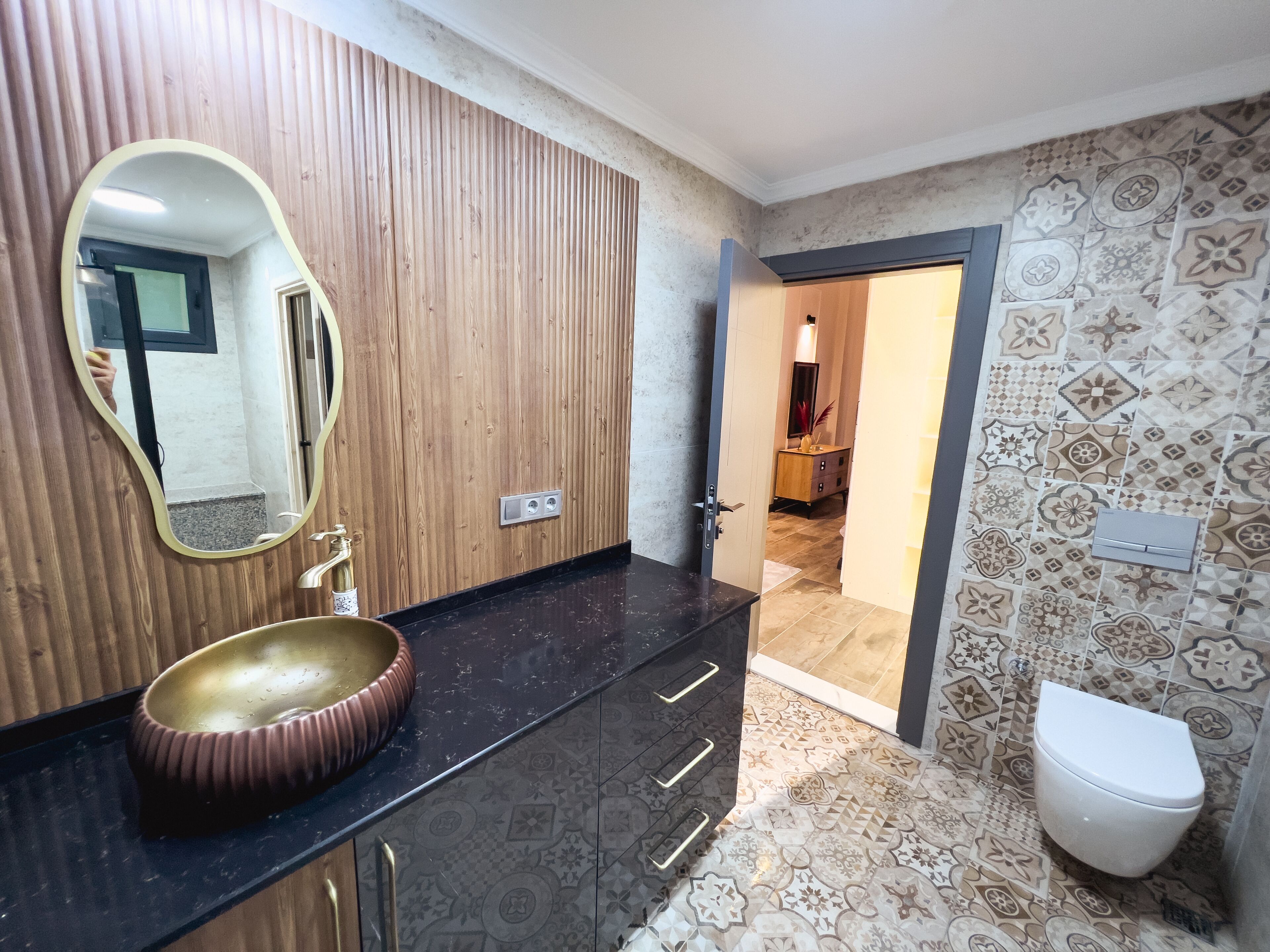 A bathroom with a wooden vanity, a round sink, a mirror, a toilet, and patterned tiles.