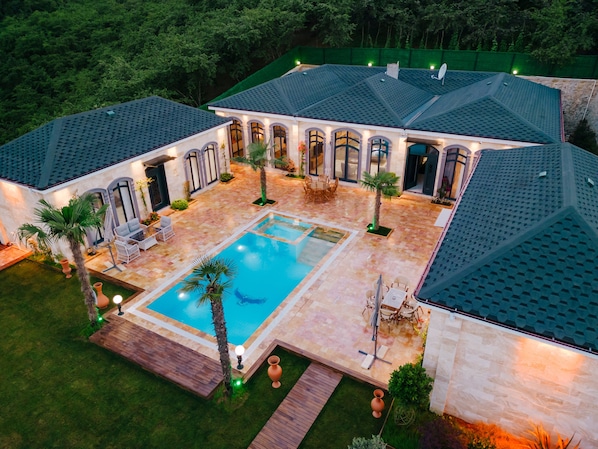 A large, well-lit outdoor pool area with a tiled deck, surrounded by a landscaped garden and a building with a tiled roof. - Arborea Hotel (Yomra)
