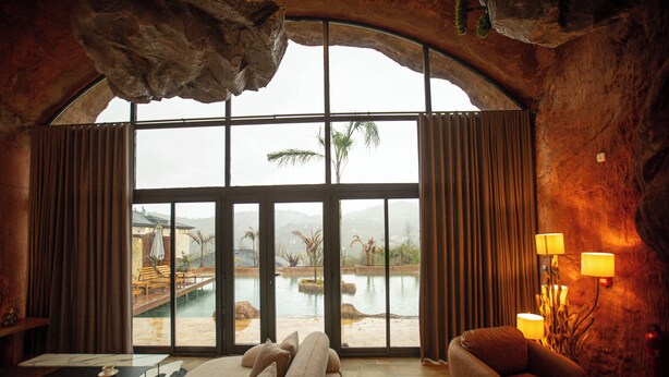 Une pièce spacieuse dotée d’une grande fenêtre en arc d’où l’on peut admirer une piscine et des palmiers, ainsi que d’un canapé confortable et d’une lampe.