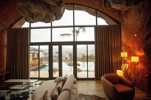 Une pièce spacieuse dotée d’une grande fenêtre en arc d’où l’on peut admirer une piscine et des palmiers, ainsi que d’un canapé confortable et d’une lampe.