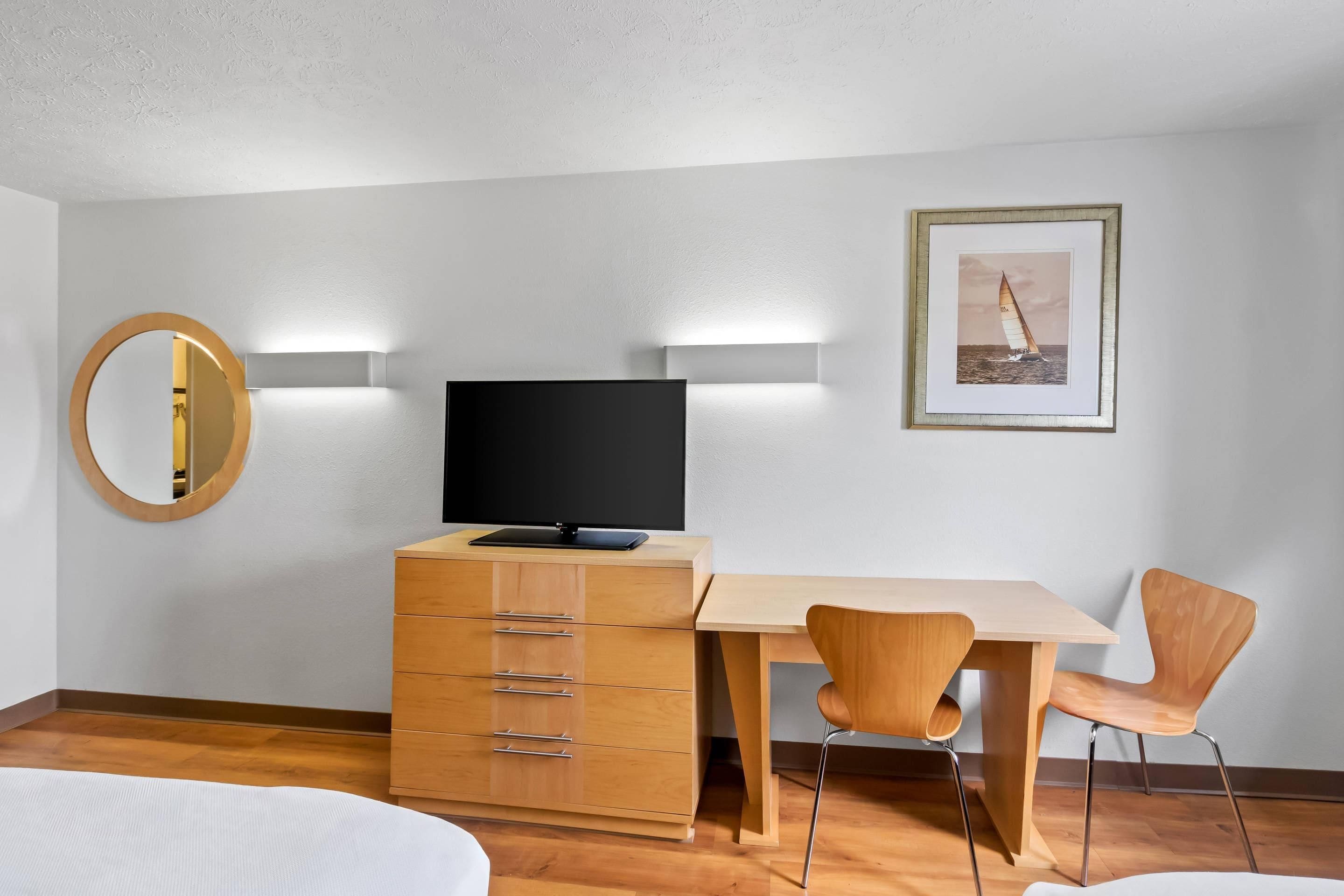 A hotel room with a bed, a TV on a wooden dresser, a desk with a chair, a framed picture on the wall, and a round mirror.