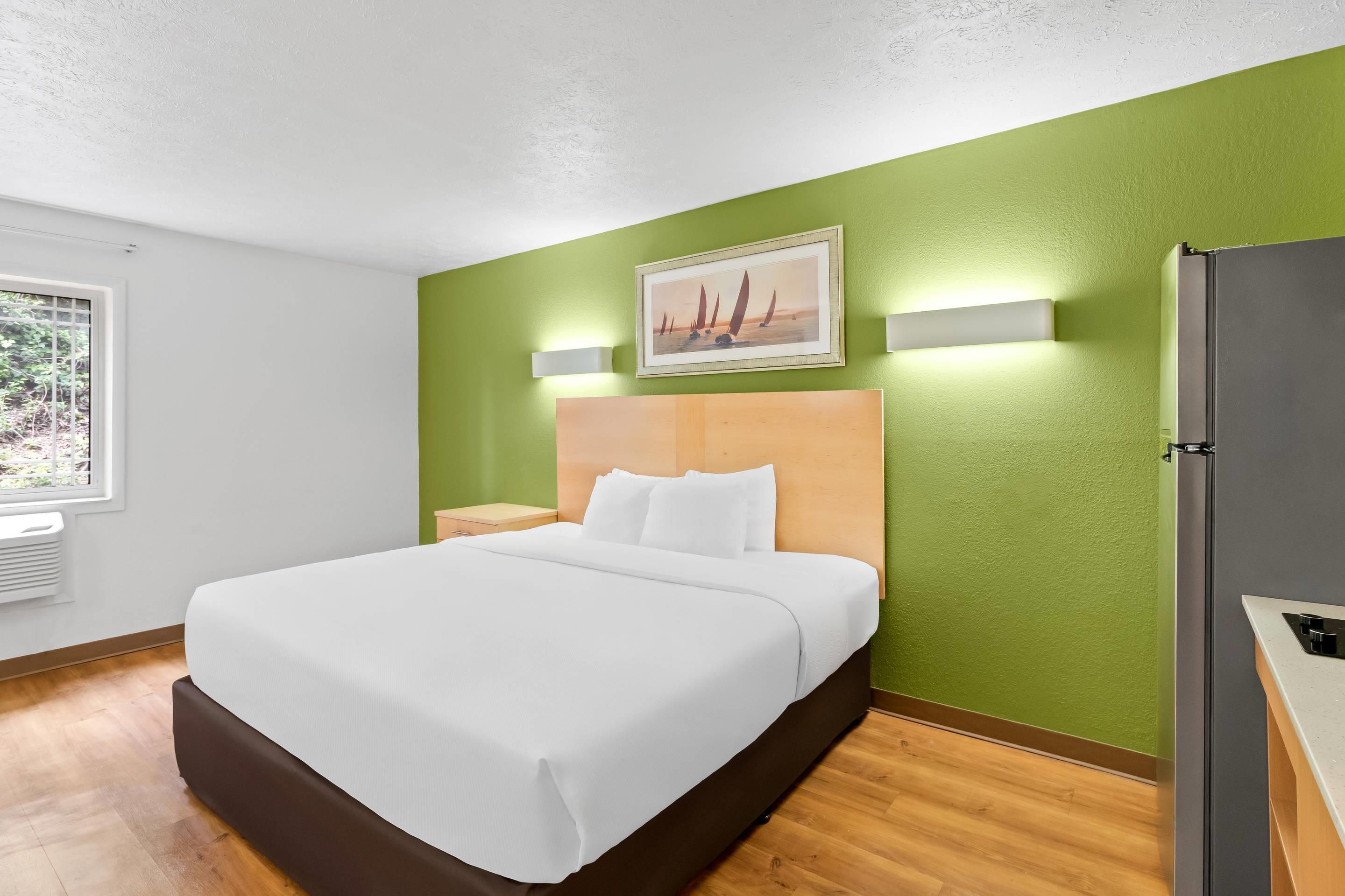 Quarto de hotel com cama, mesa de cabeceira, luminária na parede, quadro emoldurado e geladeira.