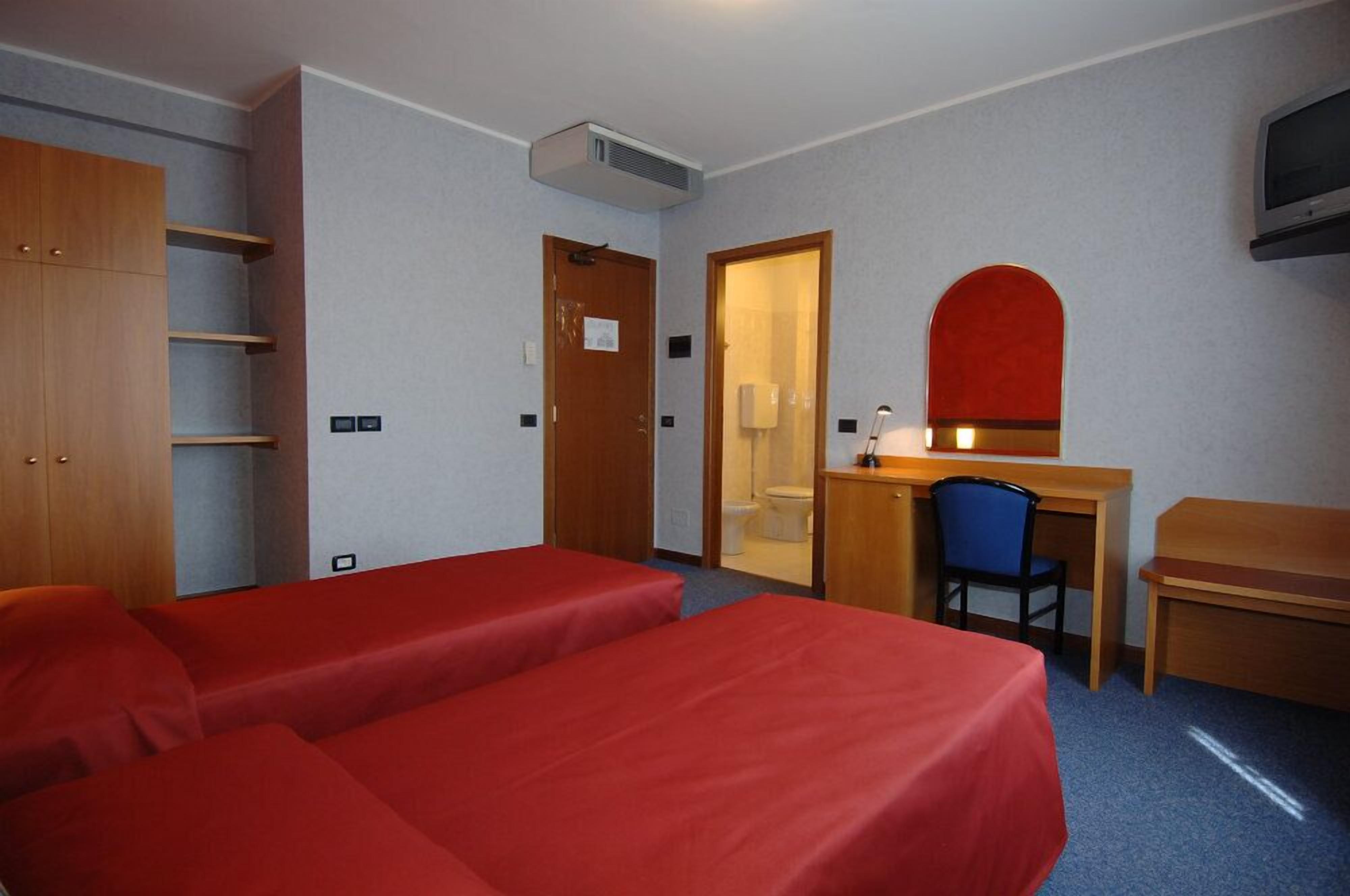Habitación de hotel con dos camas individuales, un escritorio con silla, un armario de madera y un baño visible a través de una puerta abierta.