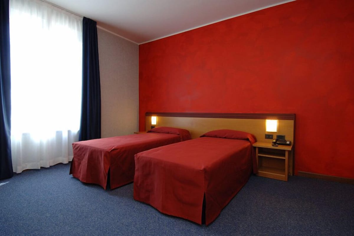 A hotel room with two single beds, a nightstand with a lamp, and a window with curtains.