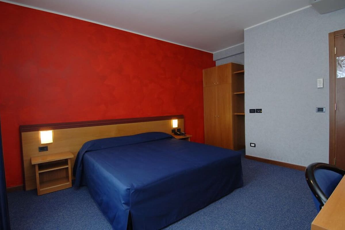 A hotel room with a single bed, a wooden wardrobe, a desk with a chair, and a nightstand with a lamp.
