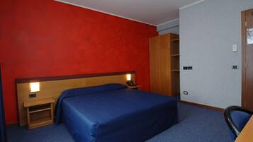 Camera d'albergo con un letto singolo, un'armadietto in legno, una scrivania con sedia e un comodino con una lampada.