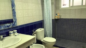Een badkamer met een wit wastafelmeubel, twee toiletten, een bidet en een douchegedeelte met een blauw douchegordijn.