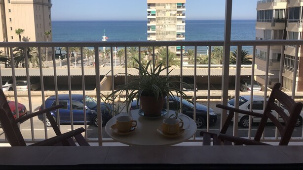 Een balkon met een tafel en stoelen, met uitzicht op de kust, gebouwen en geparkeerde auto's.