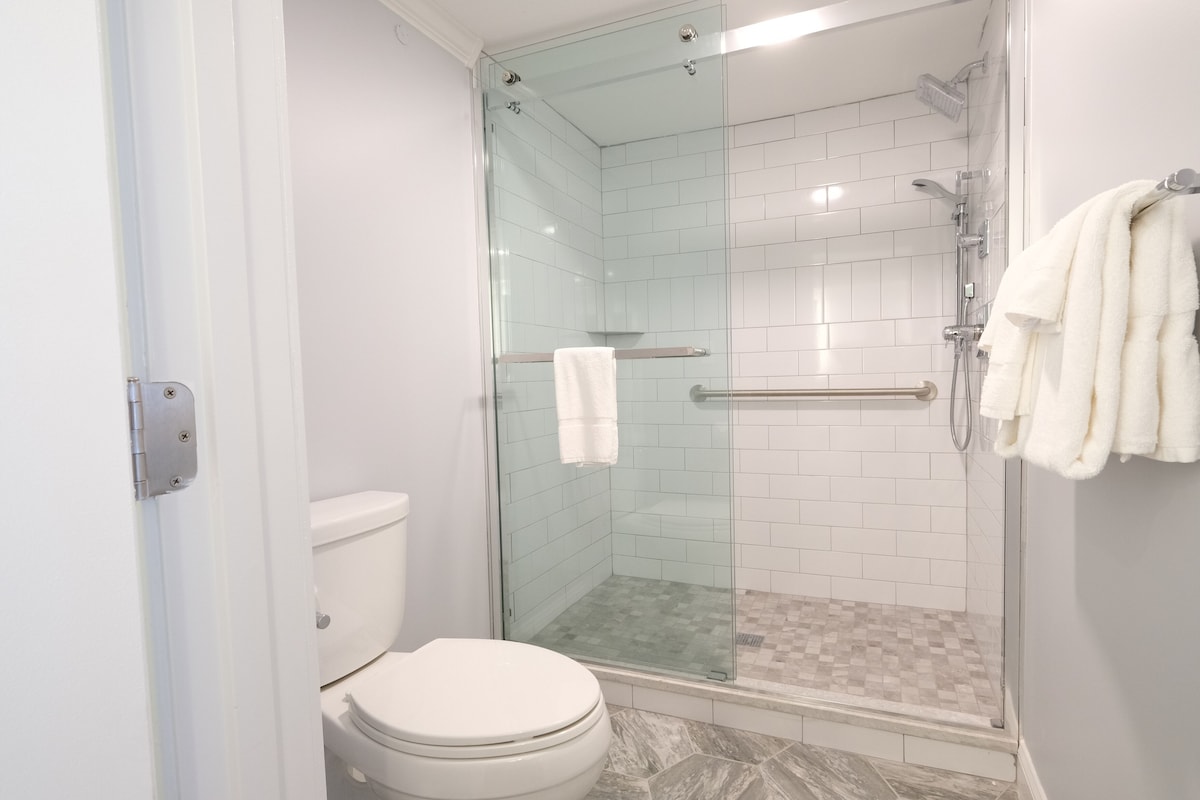 A modern bathroom with a glass-enclosed shower, white tiles, a toilet, and a towel rack with towels.