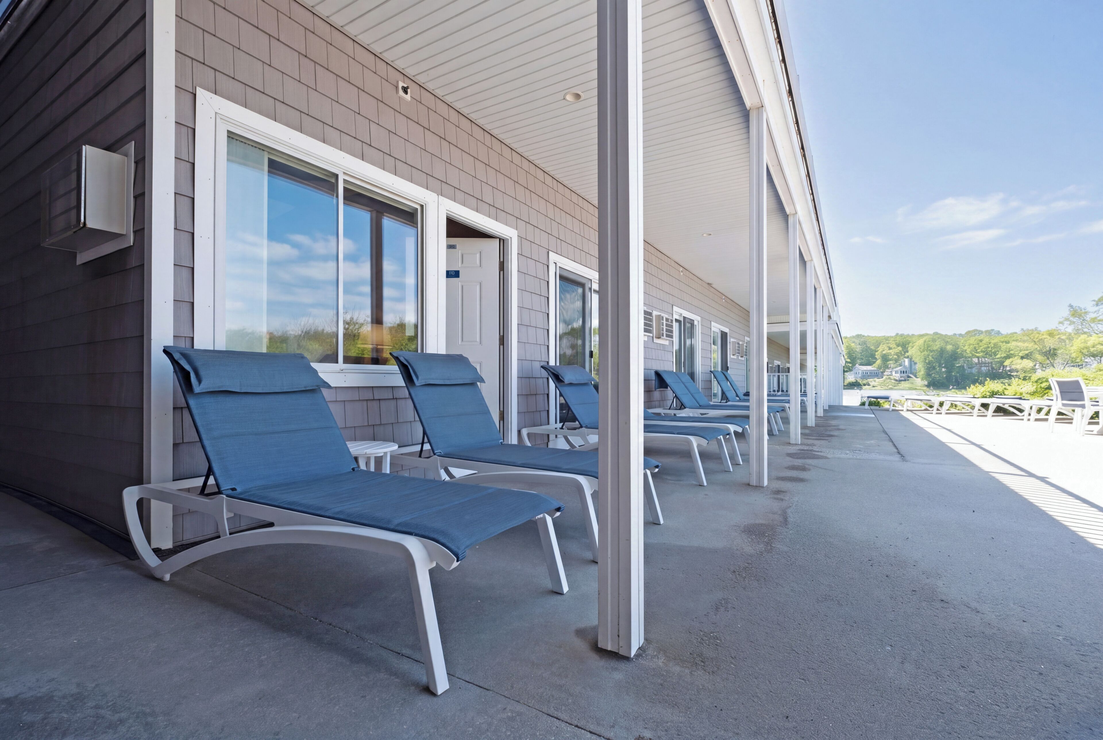A covered outdoor area with lounge chairs aligned along a building's exterior wall, featuring large windows and a clear view of the surrounding landscape.