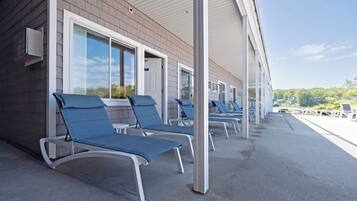 A covered outdoor area with lounge chairs aligned along a building's exterior wall, featuring large windows and a clear view of the surrounding landscape.
