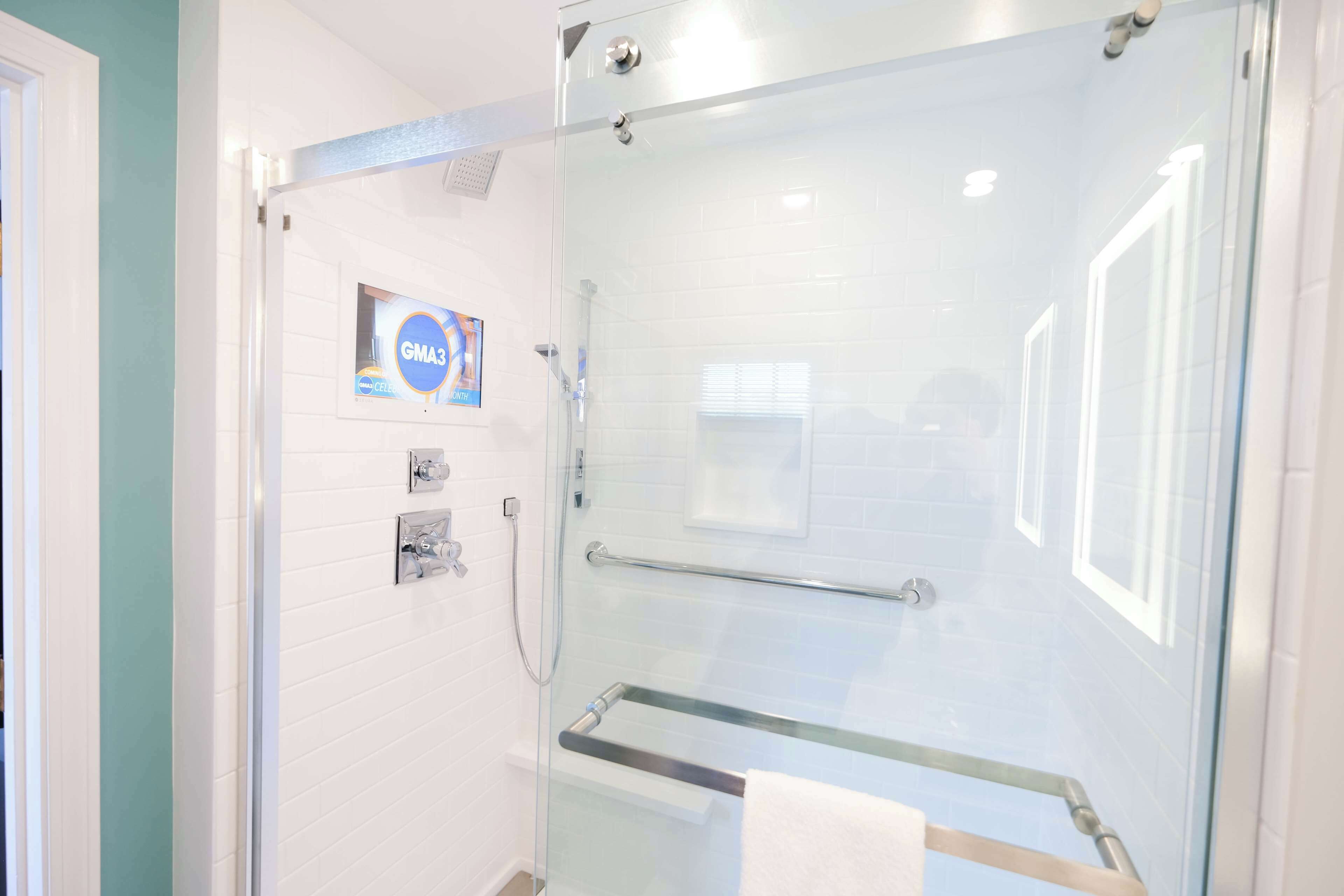 A glass-enclosed shower with a glass door, a towel rack, and a GMA3 sign on the wall.