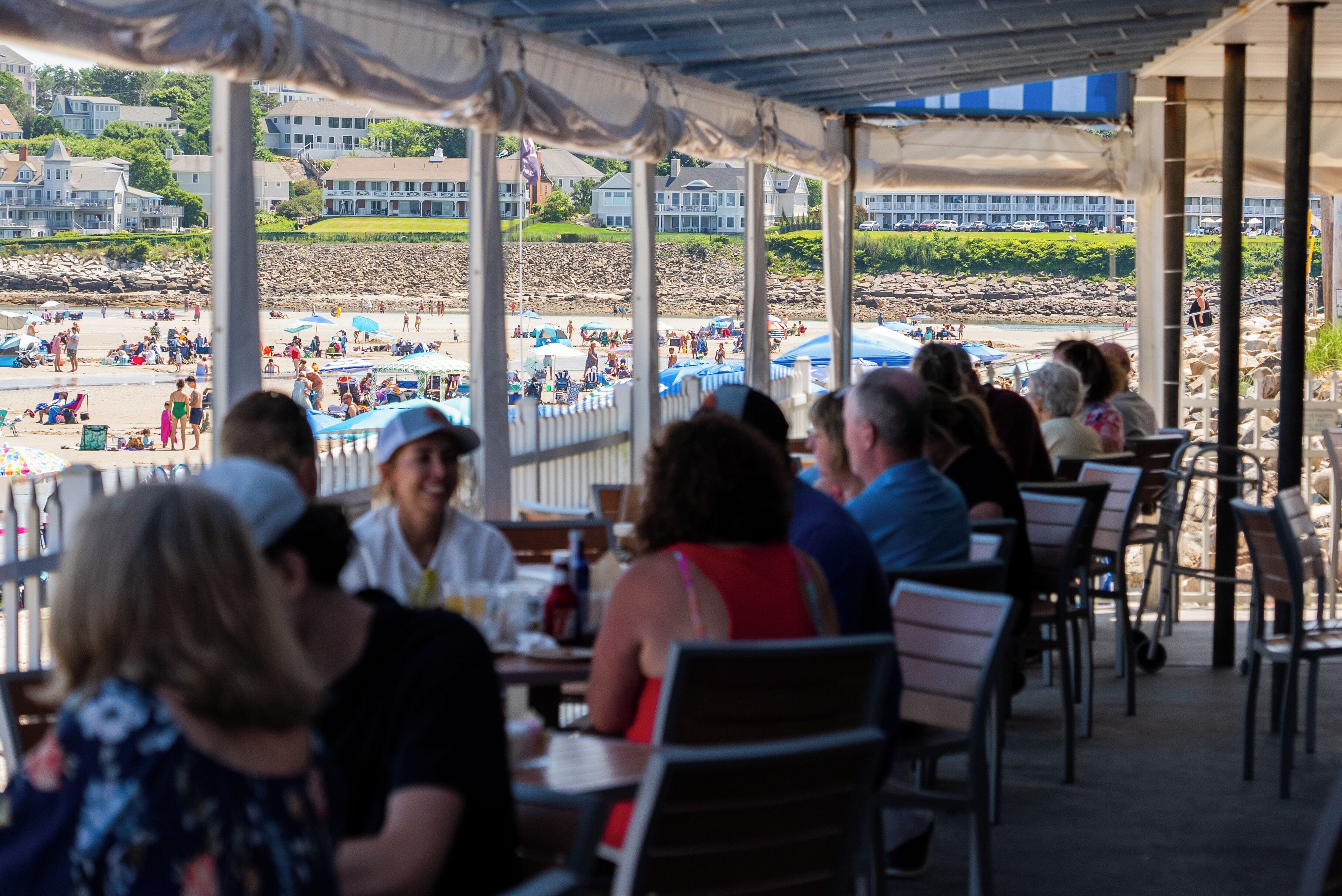 A covered outdoor seating area with tables and chairs, overlooking a beach with people and umbrellas.