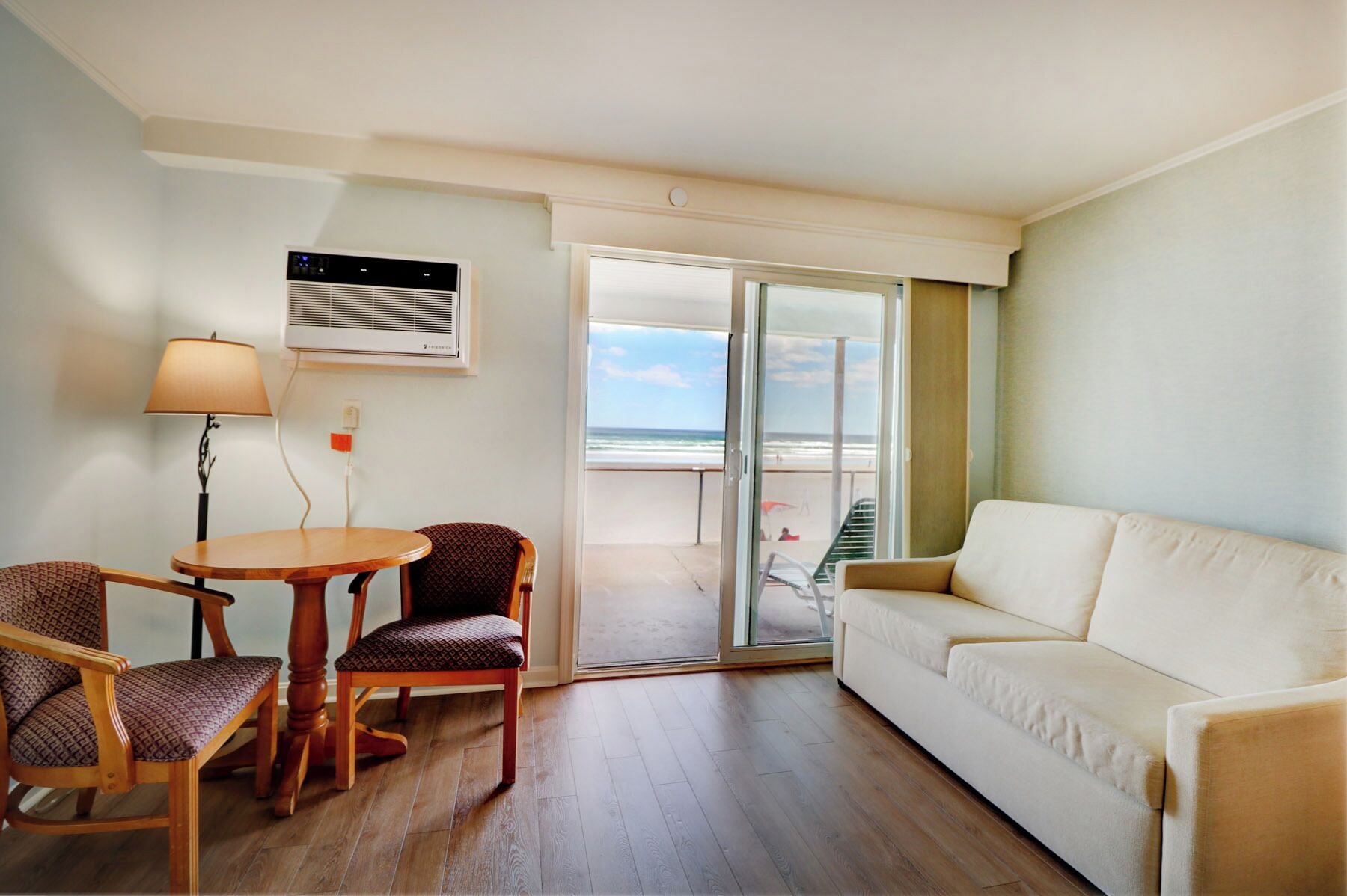 Een kamer met een houten vloer, een witte bank, een eettafel met twee stoelen, een lamp en een groot raam met uitzicht op het strand.
