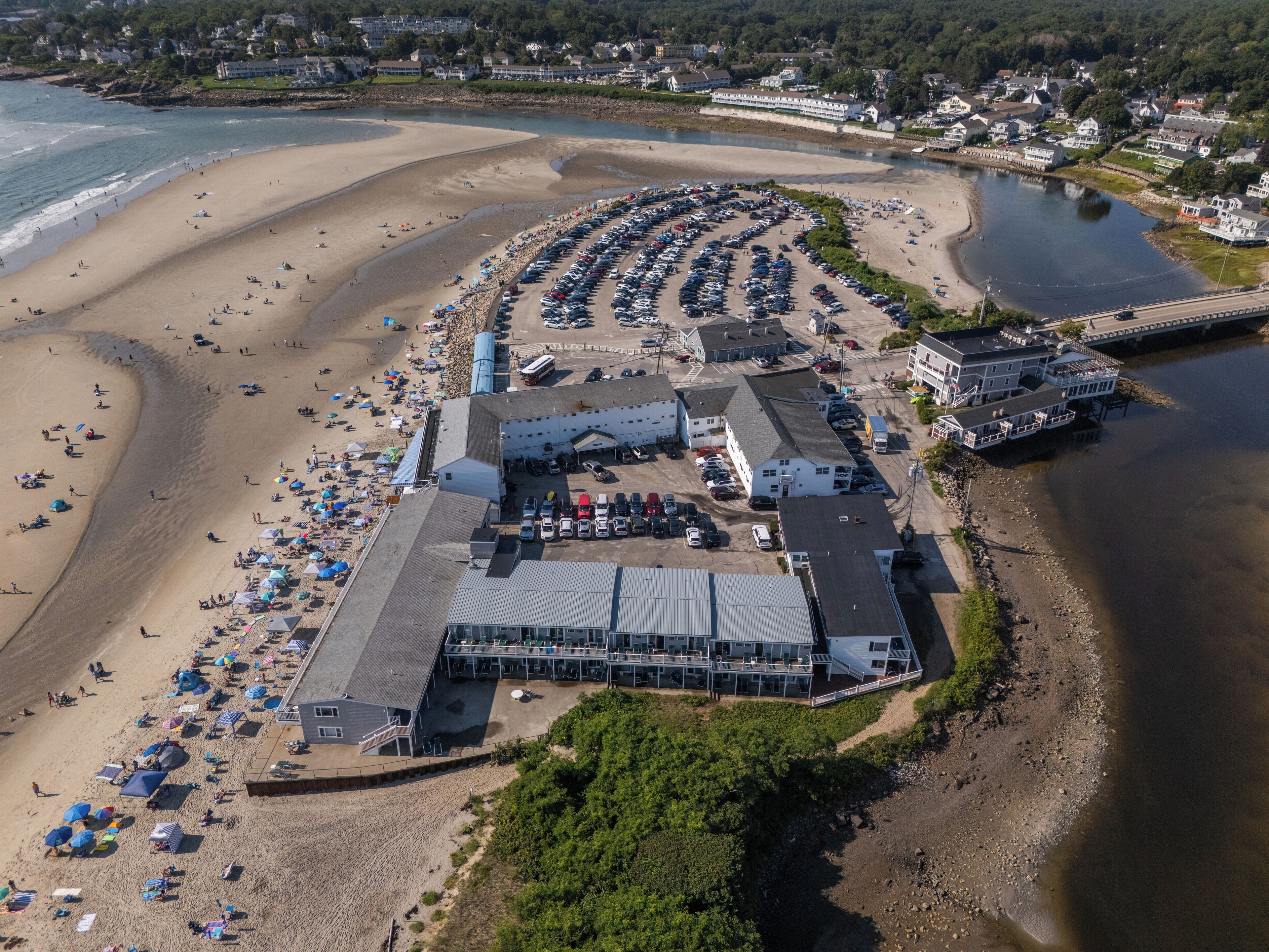 Luchtbeeld van een hotel aan het strand met een grote parkeerplaats, strandparasols en een gebouw met meerdere verdiepingen.