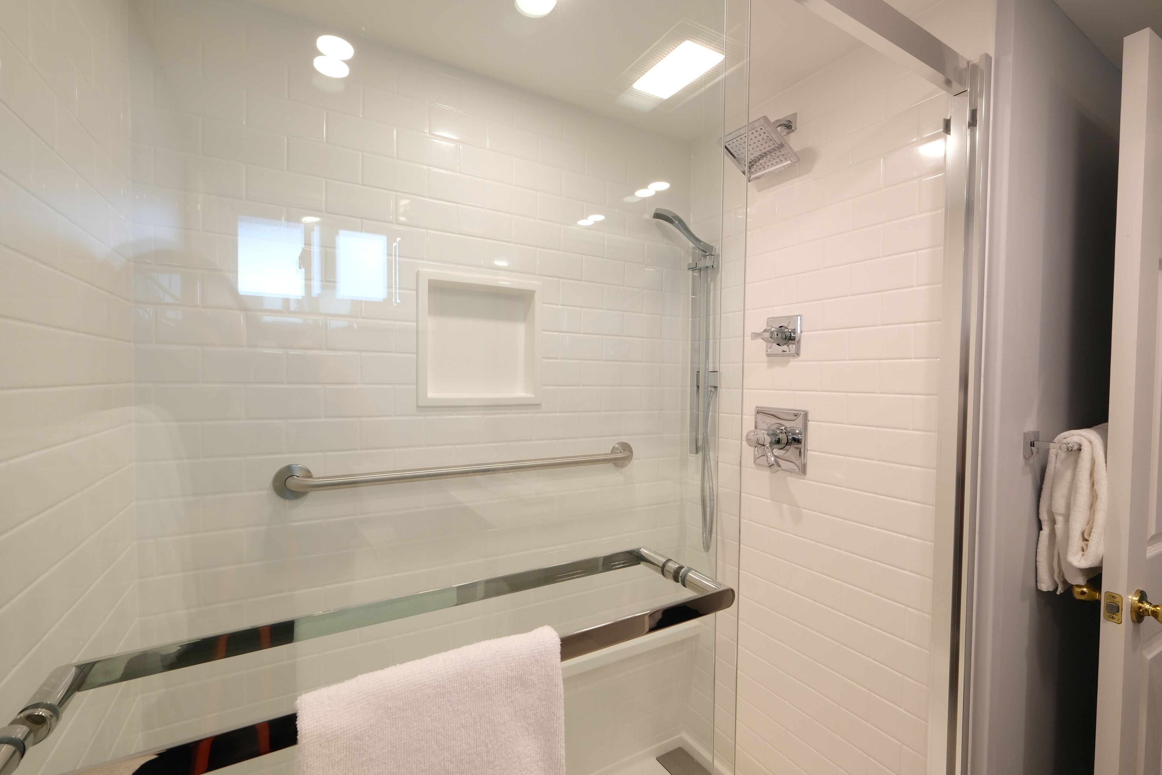 A modern bathroom with white tiled walls, a glass shower enclosure, a glass bathtub, and a white towel on the door.