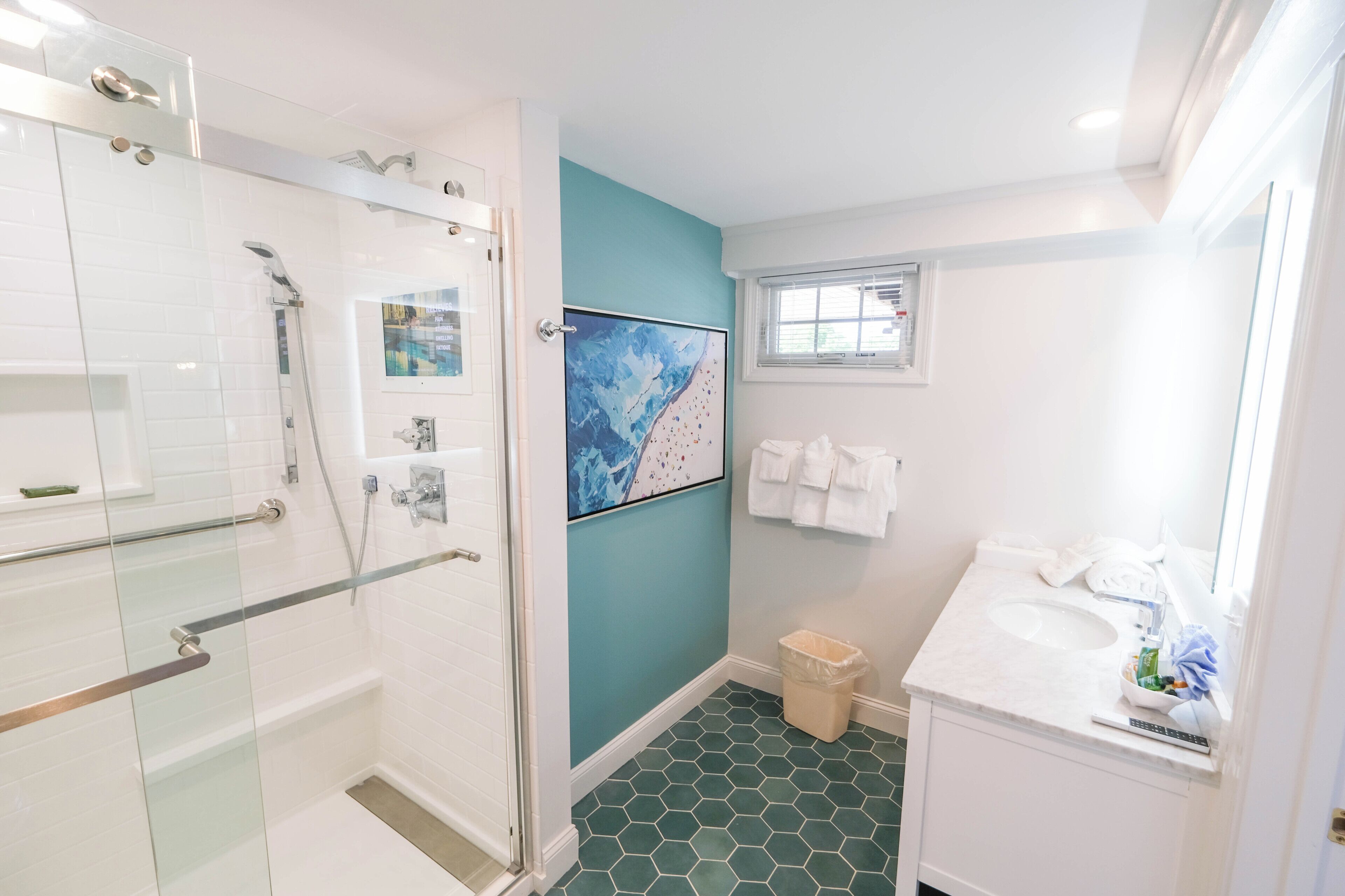 A bathroom with a glass shower enclosure, a sink with a mirror, a trash bin, and a wall-mounted towel rack.