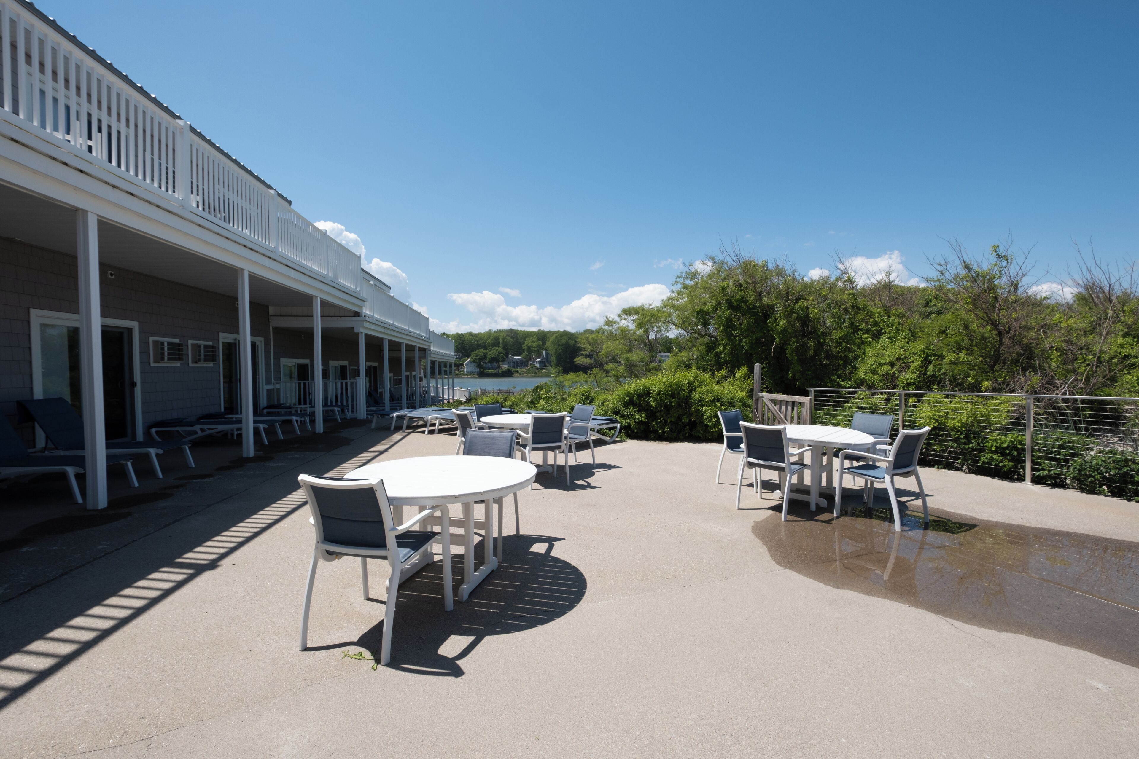 A covered outdoor patio with tables and chairs, a white building with balconies, and a view of greenery and a body of water.