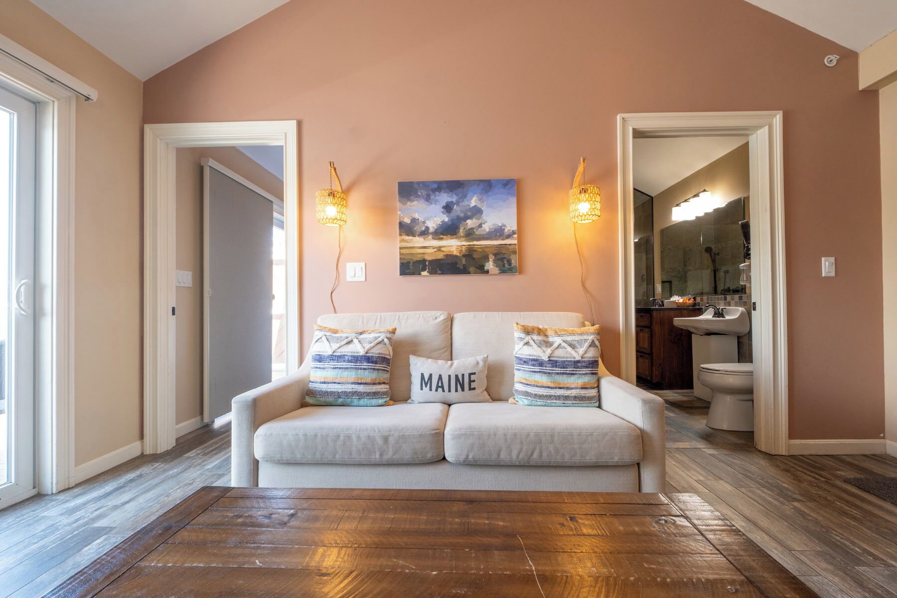 Un salon avec un canapé beige, deux lampes fixées au mur, un tableau et une vue sur la cuisine.