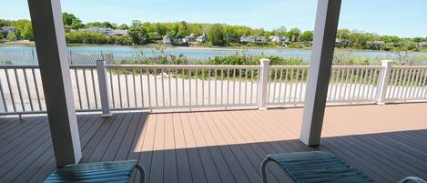 A covered deck with two green lounge chairs, a white railing, and a view of a lake with houses in the background.