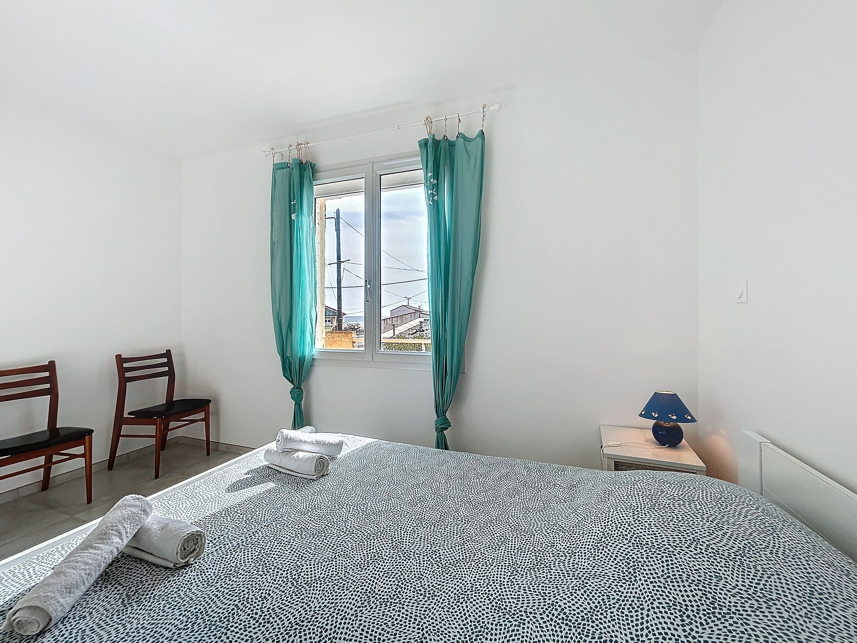 Een slaapkamer met een bed, twee stoelen, een raam met gordijnen, een lamp en een uitzicht op gebouwen.