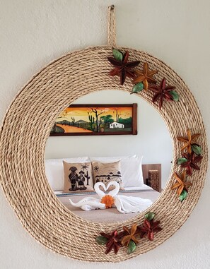 A woven circular mirror with decorative elements and a painting of a landscape in the background.