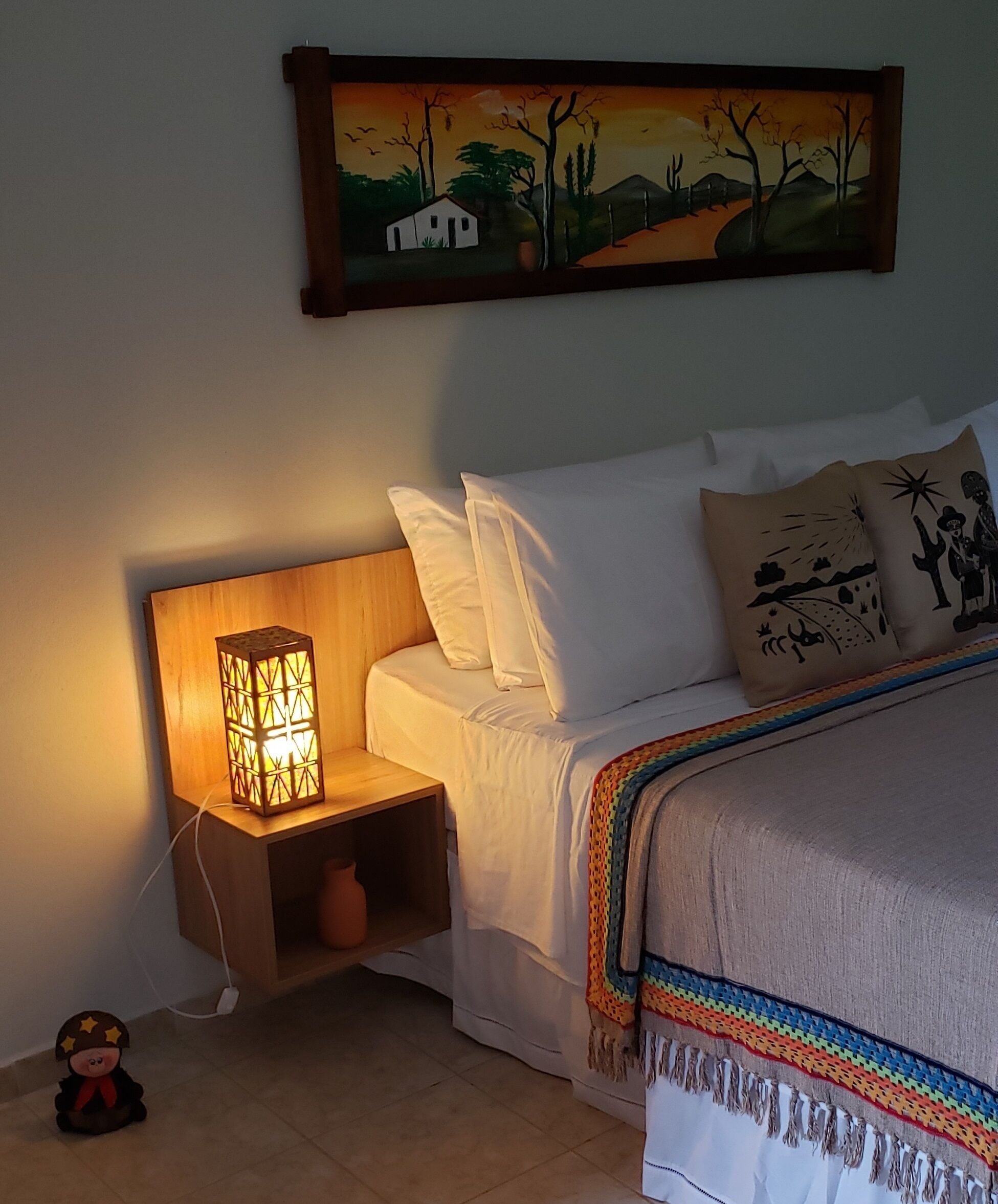 A bedroom with a bed, pillows, a nightstand with a lamp, a wall-mounted painting, and a small decorative figure.