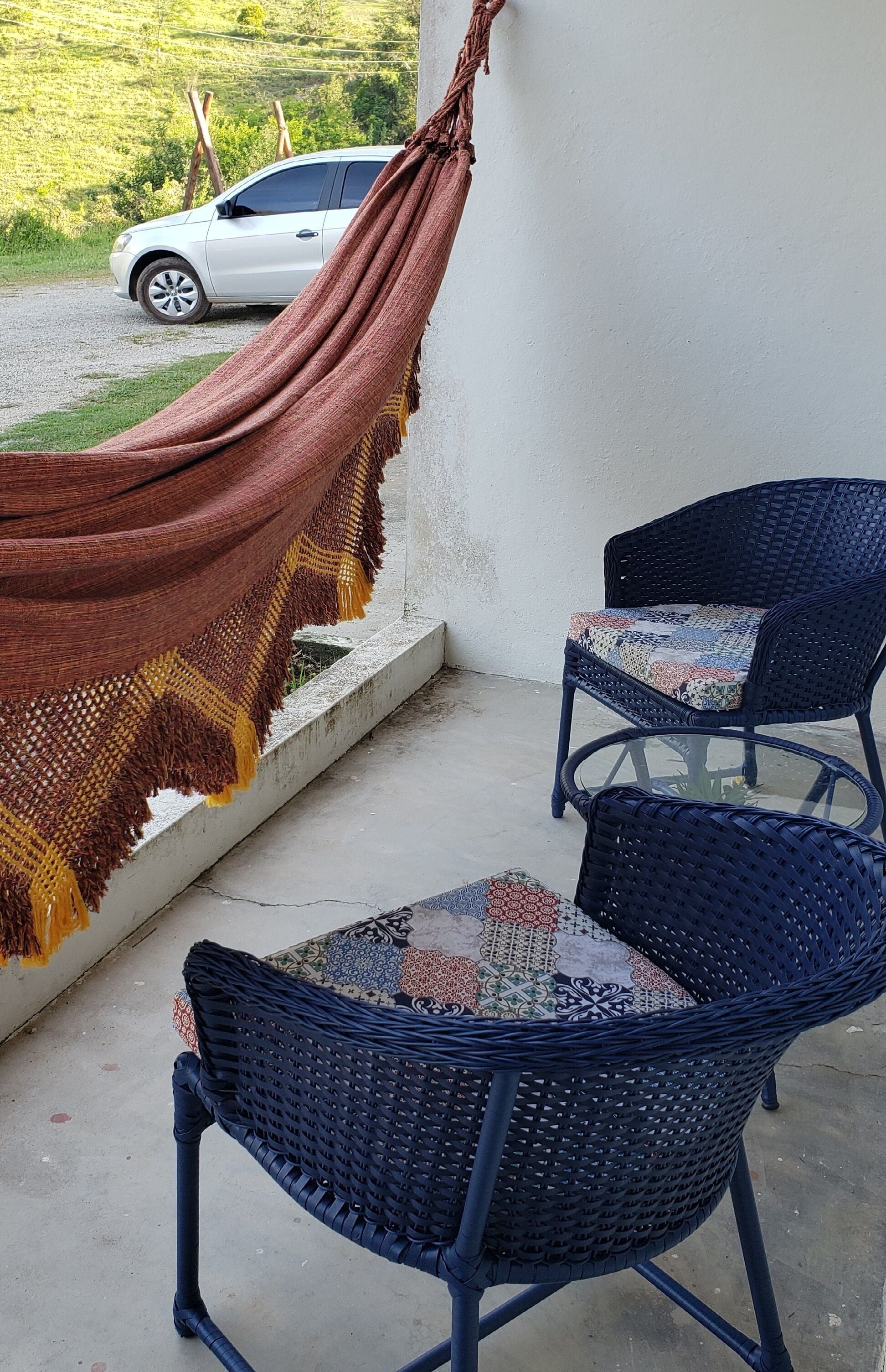 A balcony with a hammock, two wicker chairs, and a white car parked in the background.