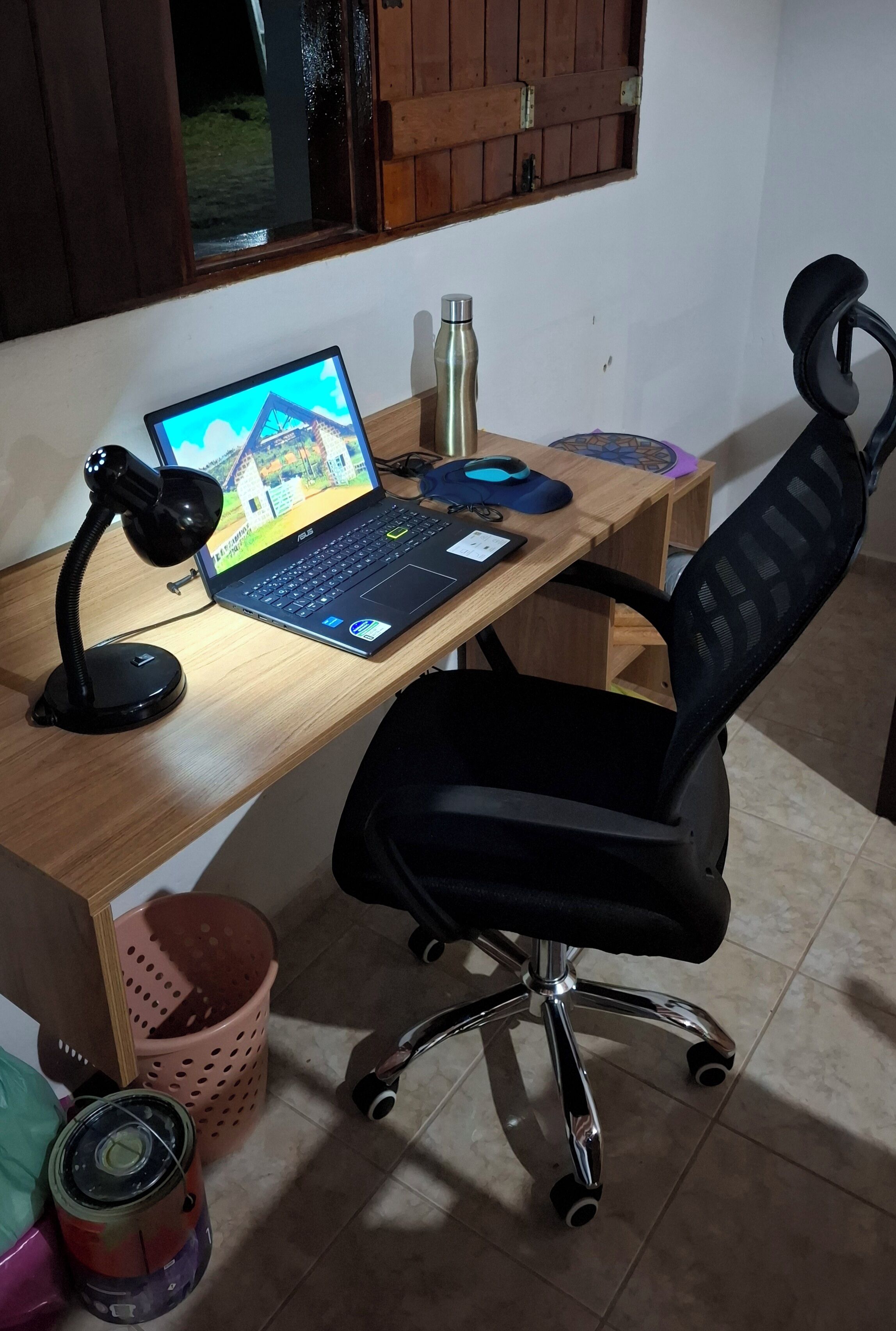 A wooden desk with a laptop, a black office chair, a pink plastic basket, and a metal water bottle.