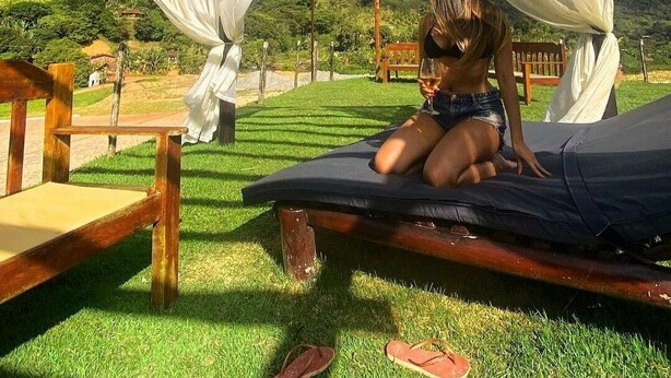 A person sitting on a hammock under a wooden structure with white fabric, surrounded by greenery and a mountain in the background.