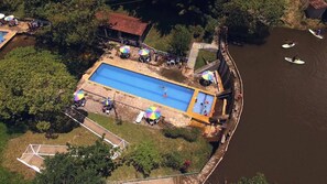 An aerial view of a swimming pool with adjacent beach areas, surrounded by trees and a river.